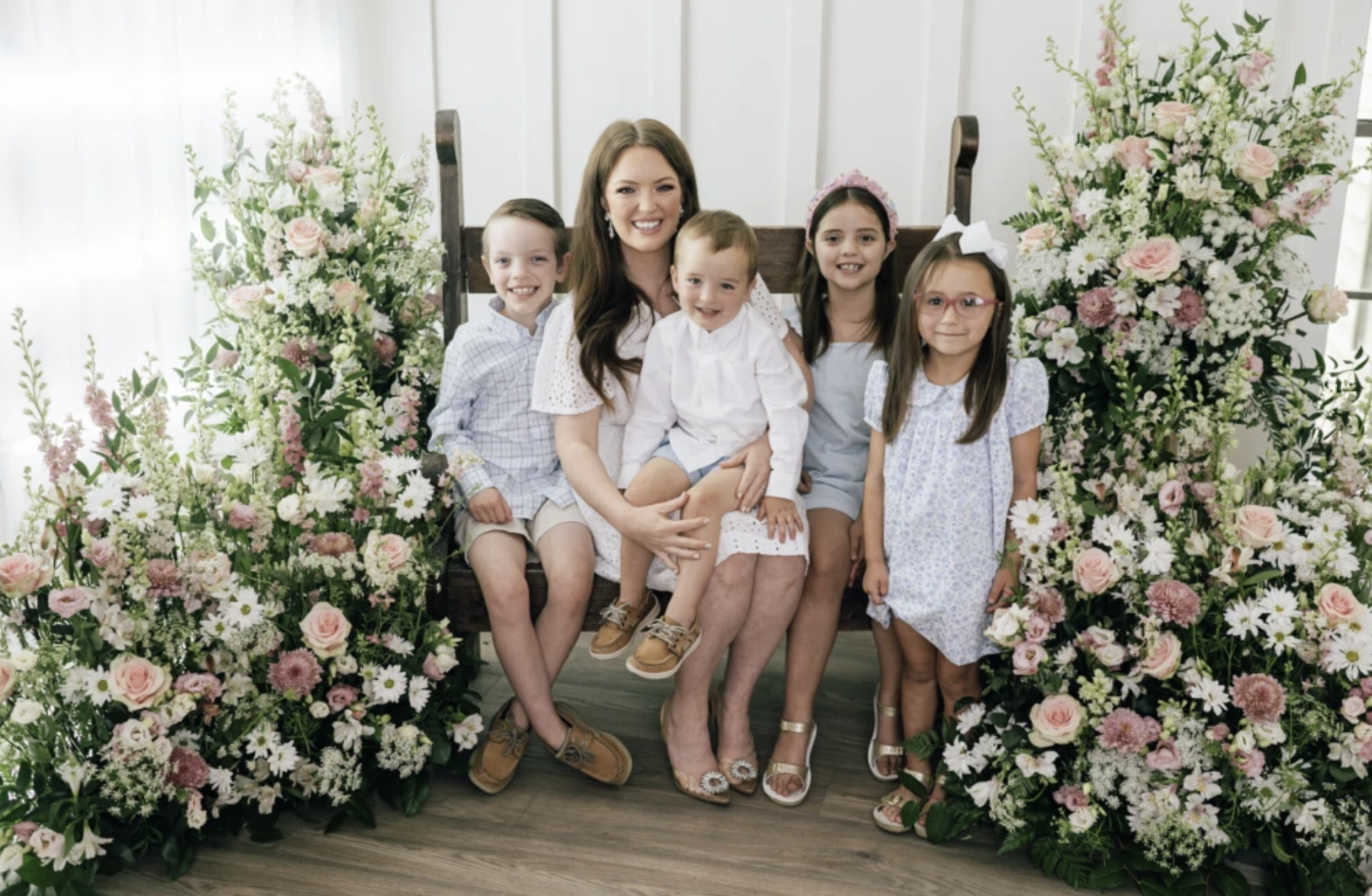 A mom and four of her children sit on a bench surrounded by flowers.